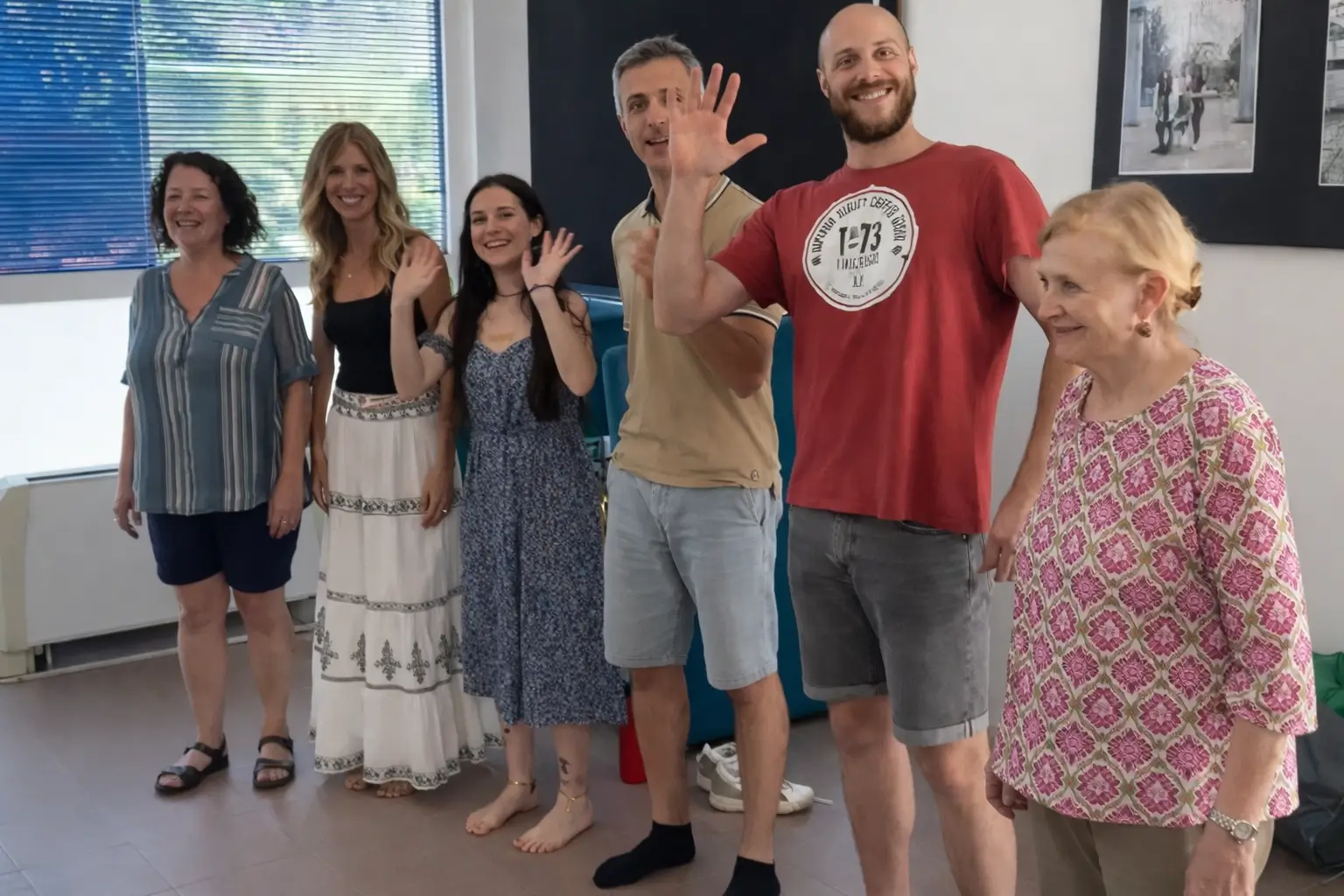 Gruppo di sei persone in piedi, sorridenti, che salutano verso la fotocamera durante un seminario in aula.